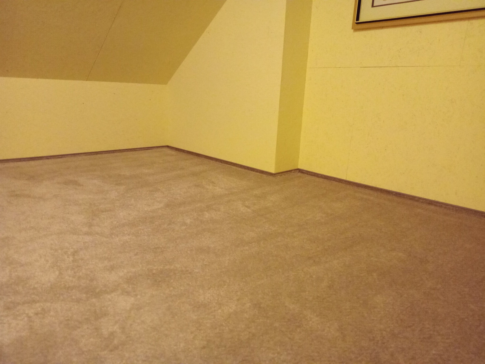 Beżowa wykładzina dywanowa w pokoju na poddaszu, widoczne listwy przypodłogowe i fragment ściany z obrazem w ramie.