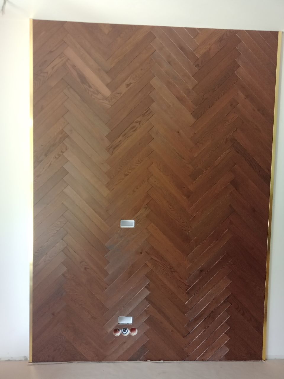 Ściana wykończona parkietem ułożonym we wzór jodełki w Wirach, z widocznymi gniazdkami elektrycznymi i ramką w kolorze złota.