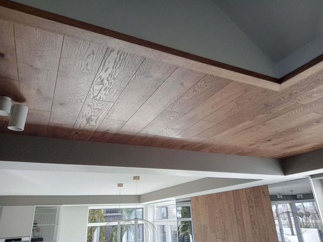 Drewniana podłoga ułożona na suficie, widok z dołu, jasne wnętrze z oknami i nowoczesnym oświetleniem w tle.