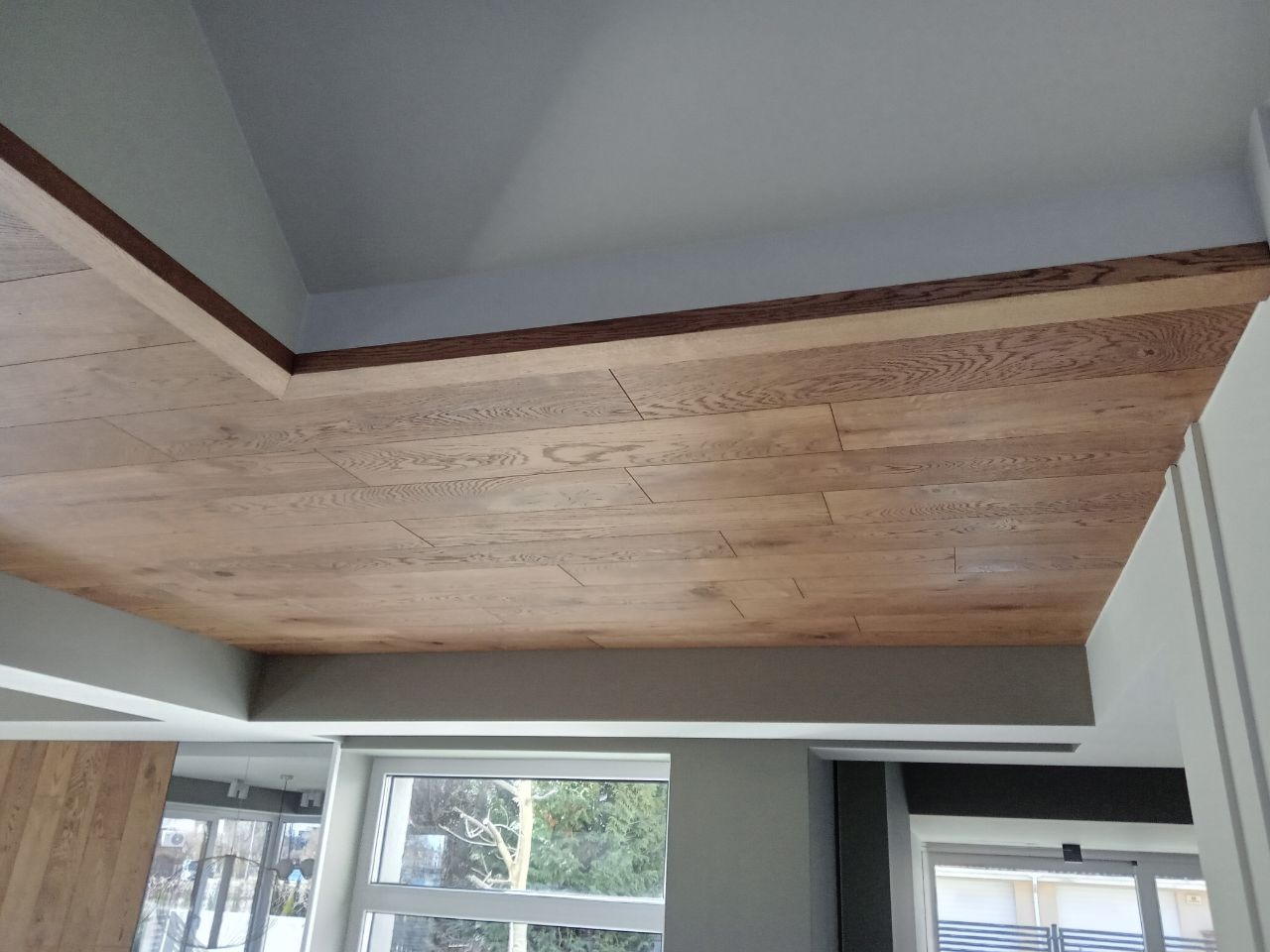 Drewniany sufit z jasnych paneli, wykończony ciemniejszą listwą, widok od dołu, okna w tle.