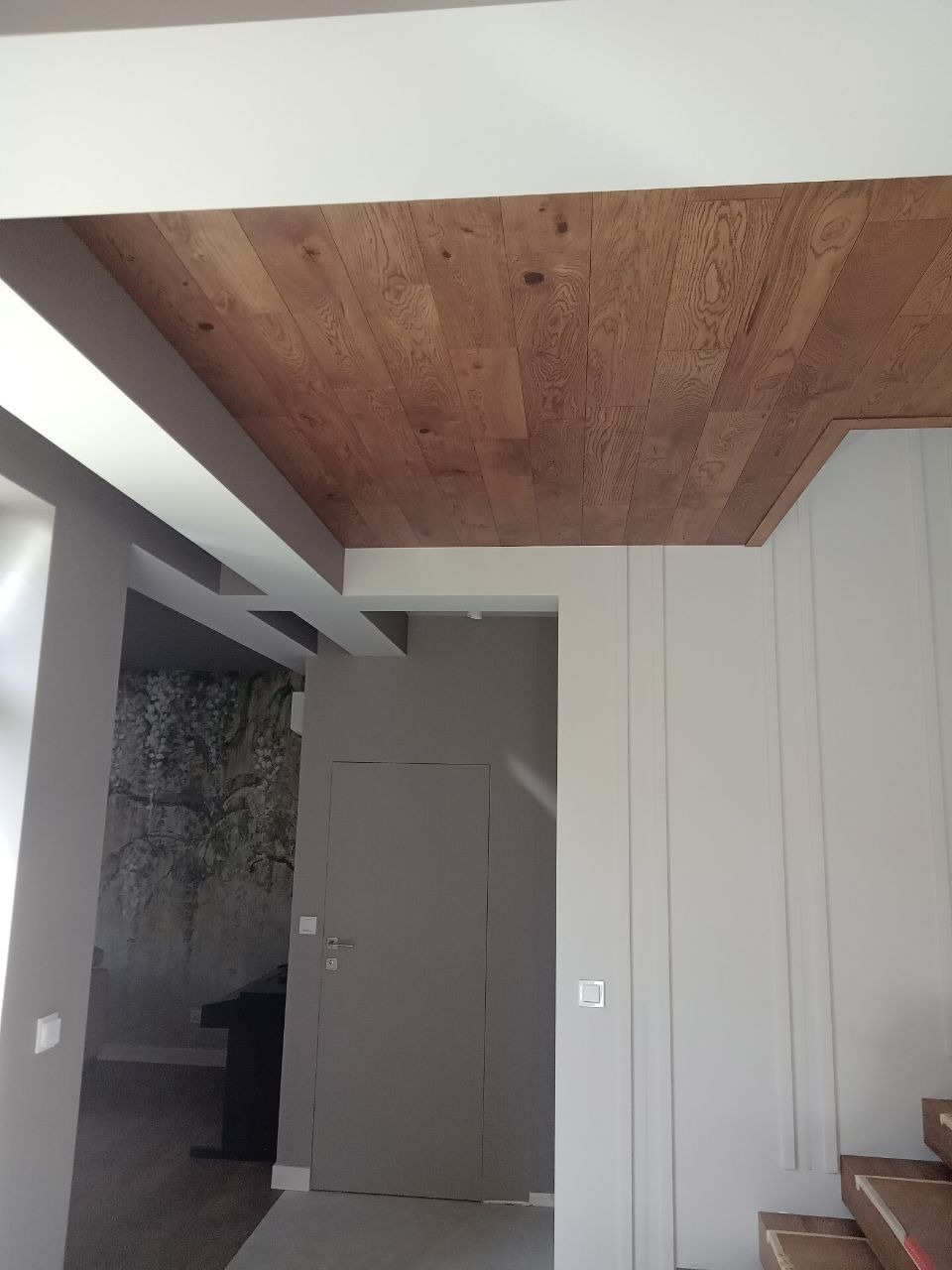Drewniana podłoga na suficie z widocznym wzorem słojów, szare ściany i białe listwy, fragment schodów w rogu.