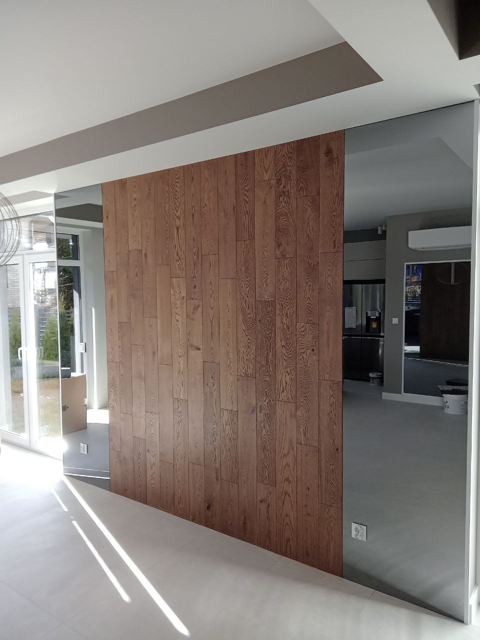 Nowoczesne wnętrze z drewnianą ścianą z paneli i lustrzanymi wstawkami tworzącymi iluzję przestrzeni, widok na salon z kuchnią w tle.