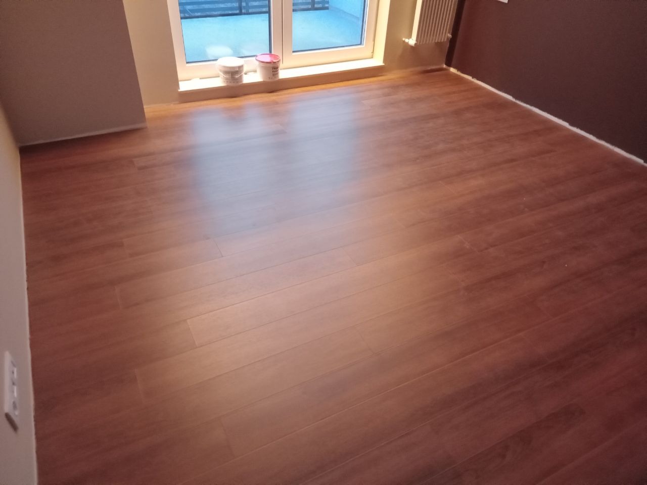 Ułożony parkiet o ciepłym, brązowym odcieniu w pustym pokoju, widoczne puszki farby przy oknie, ściany w odcieniach szarości i brązu, grzejnik na ścianie.
