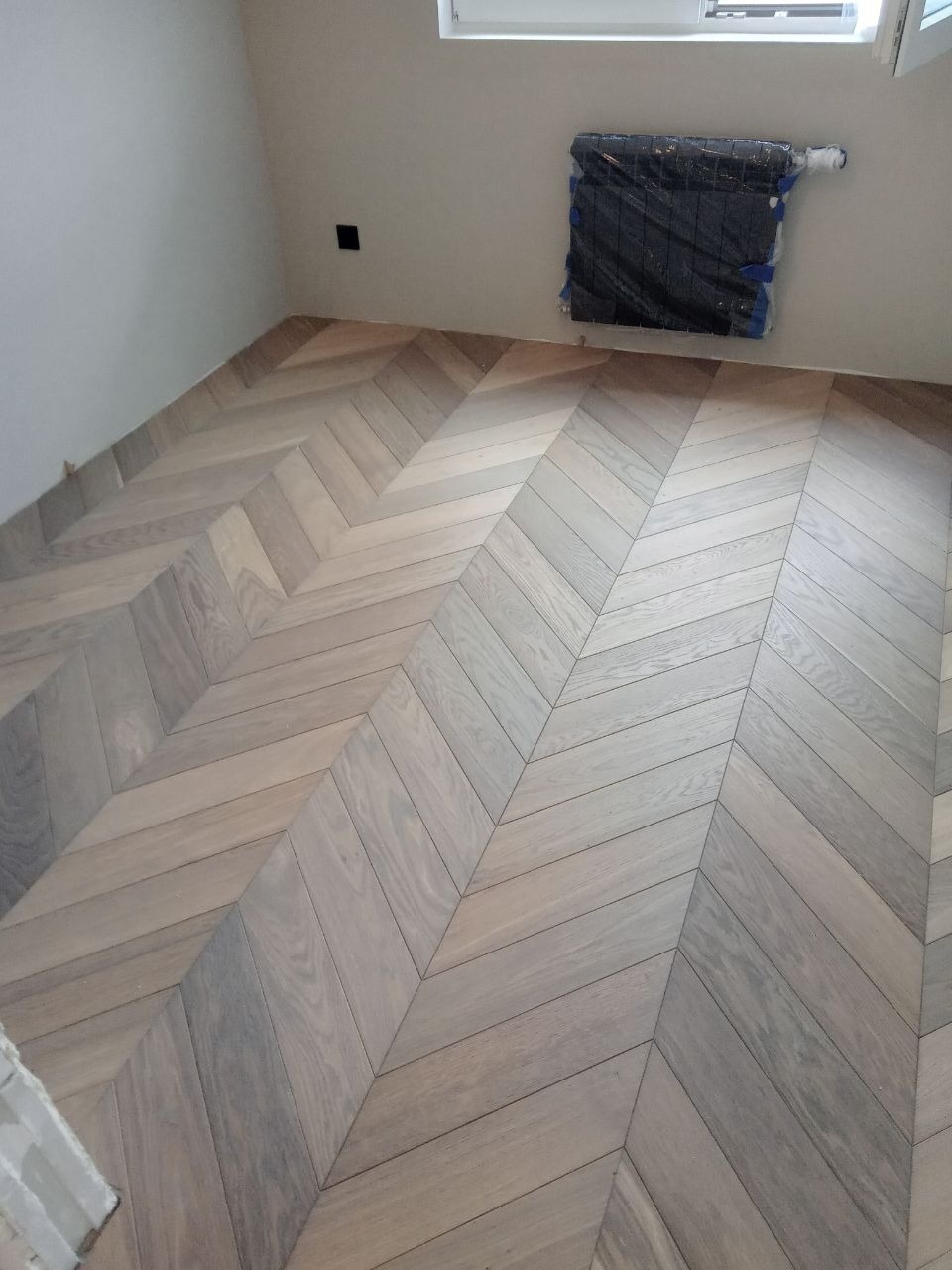 Jasny parkiet ułożony we wzór jodełki francuskiej w nowym wnętrzu, grzejnik zabezpieczony folią malarską.