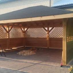 Konstrukcja drewnianej altany z czarnym dachem z gontu bitumicznego, z widoczną drabinką i stertą odpadów drewnianych wewnątrz, na tle budynku z białej cegły.