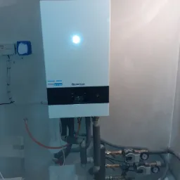 Nowy, biały piec gazowy Buderus zamontowany na surowej, szarej ścianie, widoczne rury instalacji grzewczej i pompy obiegowe.