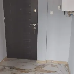 Ułożone płytki podłogowe imitujące marmur w przedpokoju, widoczna ciemna, metalowa drzwi wejściowe, biały bojler na ścianie i lampa sufitowa.