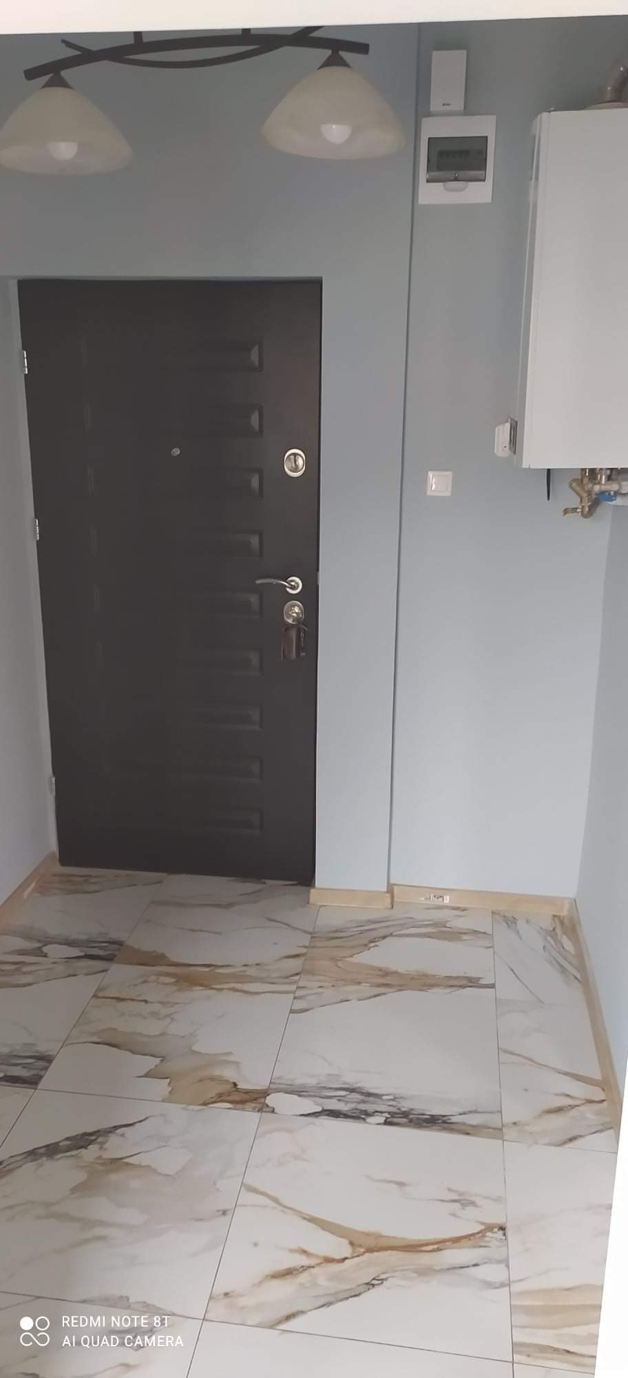 Ułożone płytki podłogowe imitujące marmur w przedpokoju, widoczna ciemna, metalowa drzwi wejściowe, biały bojler na ścianie i lampa sufitowa.