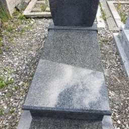 Szary, granitowy pomnik z połyskującą płytą nagrobną, otoczony drobnymi kamykami i zielenią, widoczne elementy betonowego ogrodzenia.