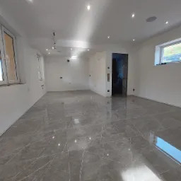 Duże, puste pomieszczenie z szarą, marmurkową podłogą z płytek, białymi ścianami, kilkoma oknami i wpuszczanymi lampami sufitowymi, widoczne niedokończone prace wykończeniowe.