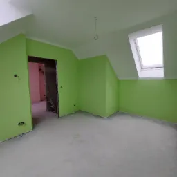 Pomalowane na jasnozielono poddasze z oknem dachowym, widoczne niedokończone gniazdka elektryczne i surowa podłoga, w tle fragment innego pomieszczenia pomalowanego na różowo.