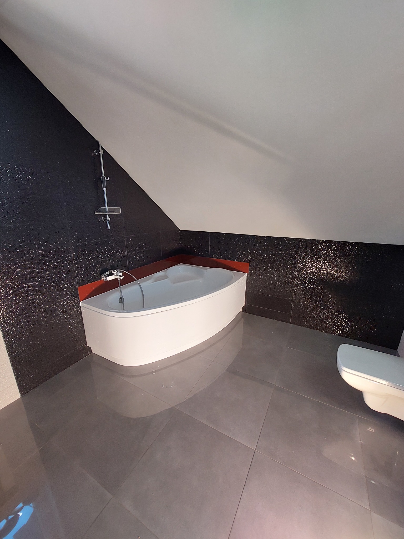 Nowoczesna łazienka na poddaszu z czarnymi, błyszczącymi płytkami na ścianach, białą narożną wanną z czerwonym akcentem oraz szarą, dużą płytką podłogową.