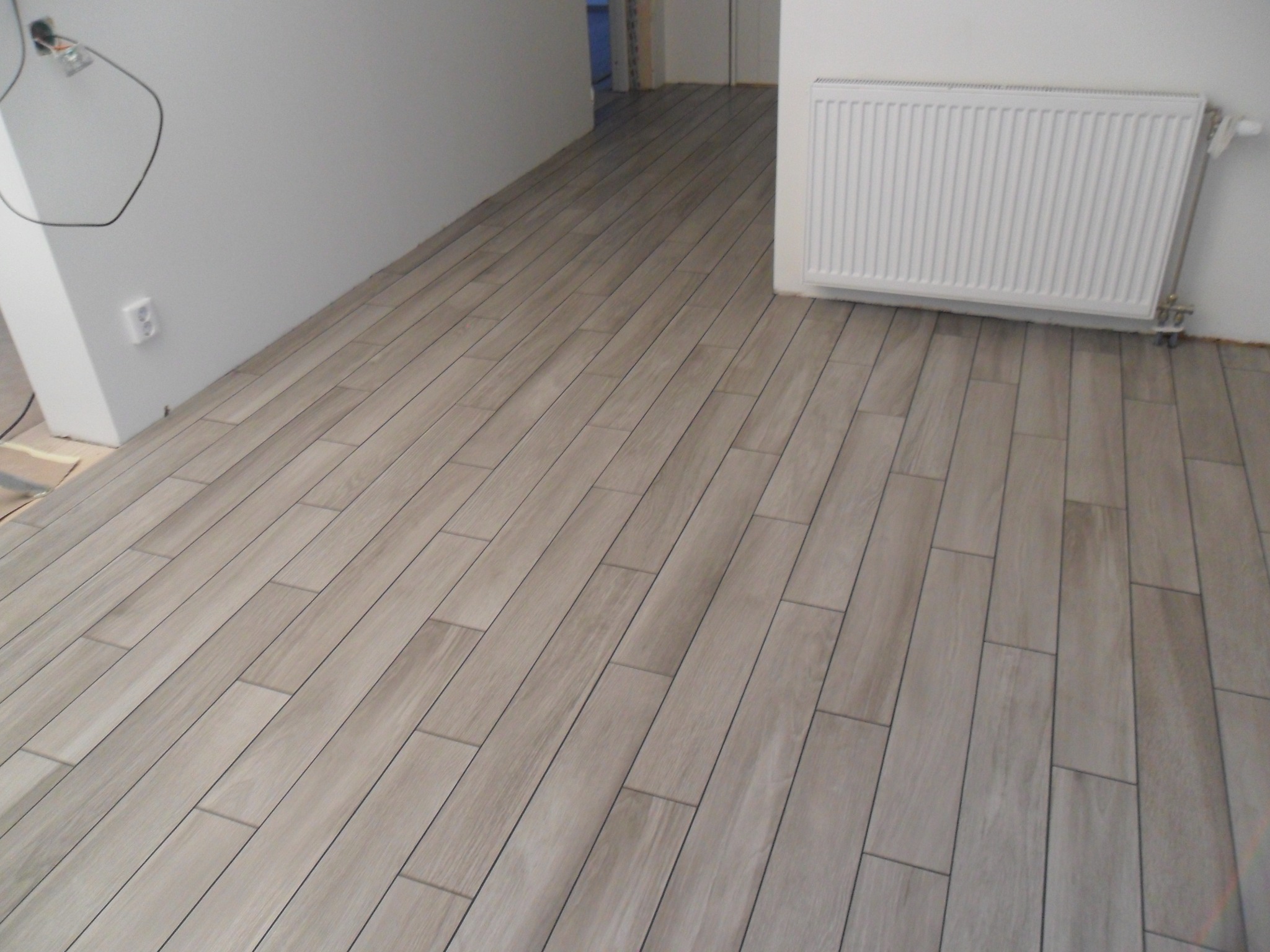 Ułożona podłoga z płytek imitujących drewno w jasnych odcieniach szarości, widoczny biały grzejnik na ścianie i wystający kabel elektryczny, ściany pomalowane na biało.