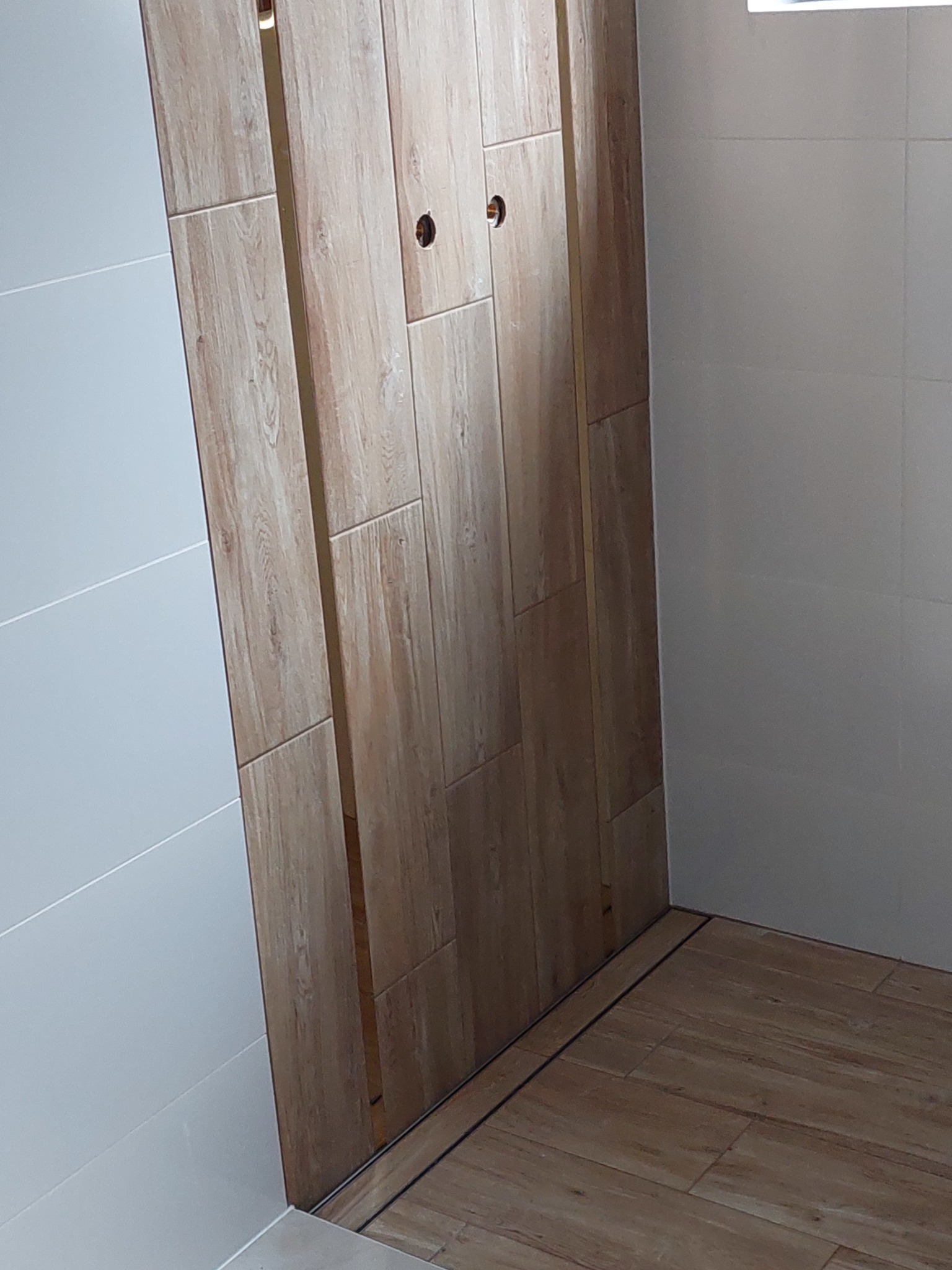 Wykończona łazienka z kabiną prysznicową typu walk-in wyłożoną płytkami imitującymi drewno, widoczne otwory na armaturę.