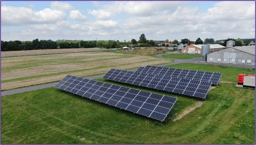 Rozległa farma z trzema rzędami paneli słonecznych na trawiastym terenie, budynki gospodarcze w tle pod pochmurnym niebem.