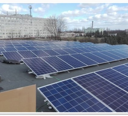 Rząd paneli słonecznych na płaskim dachu budynku, widok z góry, w tle budynek z anteną i drzewa.