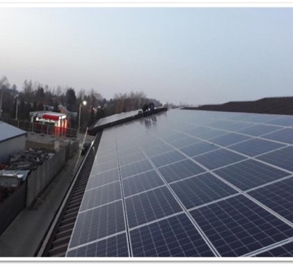 Instalacja paneli słonecznych na dachu budynku, widoczne panele ułożone w rzędy, w tle budynki i drzewa, pochmurne niebo.