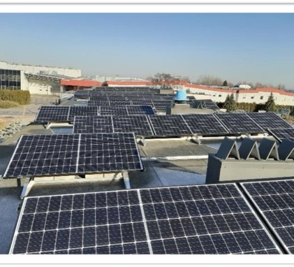 Rozległa instalacja paneli słonecznych na płaskim dachu budynku przemysłowego w słoneczny dzień. Widoczne liczne rzędy paneli, elementy wentylacyjne i fragmenty otoczenia.