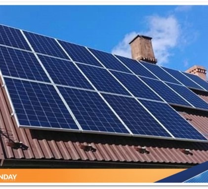 Panele słoneczne na brązowym dachu domu z ceglanym kominem, widoczne na tle błękitnego nieba.