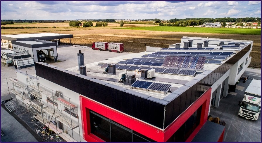 Widok z góry na budynek z panelami słonecznymi na dachu, klimatyzatorami i rusztowaniem przy ścianie. W tle pola uprawne i zabudowania.