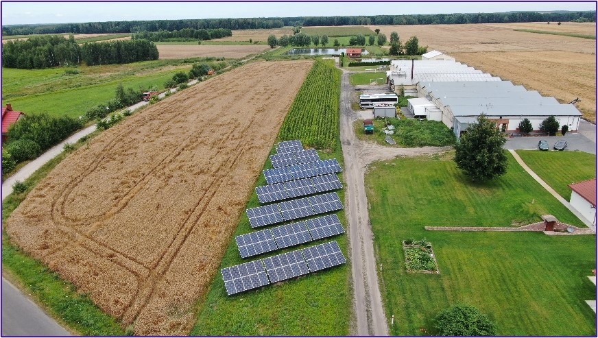 Zestaw paneli fotowoltaicznych ułożonych rzędami na skraju pola uprawnego, widok z góry, w tle budynek gospodarczy i zadrzewienia.