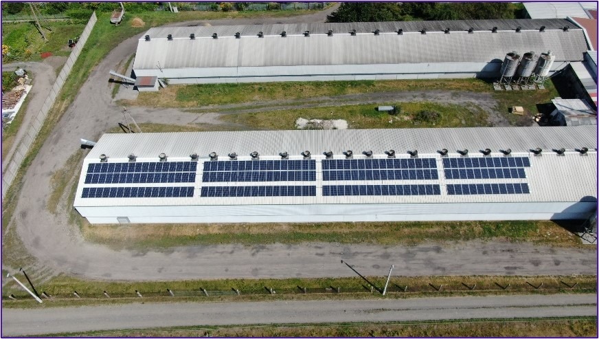 Widok z drona na duży budynek gospodarczy z zainstalowanymi panelami słonecznymi na dachu, widoczne również silosy i budynki w tle.