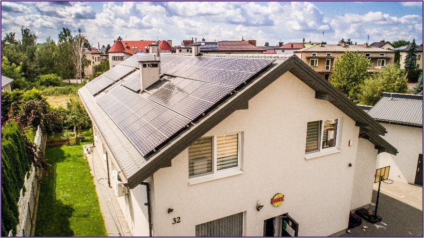 Widok z lotu ptaka na dom jednorodzinny z panelami słonecznymi na dachu, biała elewacja, szare okna i dach, widoczny trawnik i inne budynki w tle.