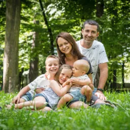 Uśmiechnięta rodzina z trójką dzieci siedzi na trawie w parku, z widocznymi drzewami w tle.