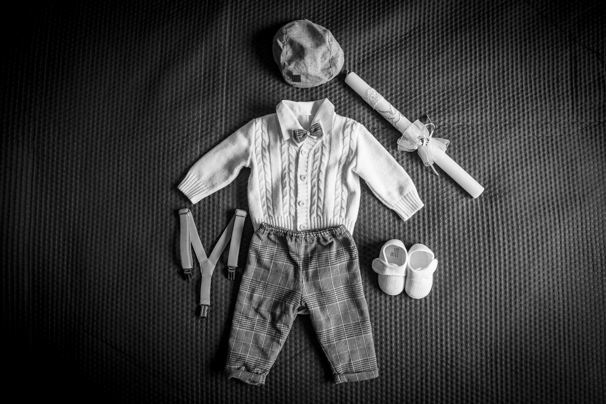 Czarno-białe ujęcie z góry prezentujące ubranka chłopięce: sweterek z muszką, kraciaste spodnie, szelki, buciki, kaszkiet oraz świecę do chrztu, ułożone na teksturowanym tle.