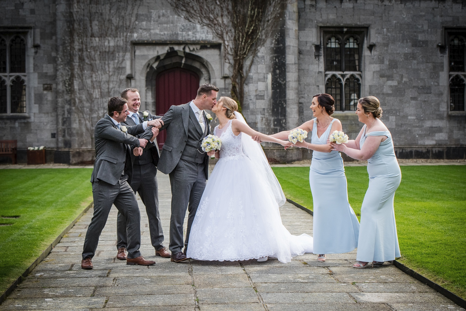 Panna młoda i pan młody całują się na tle kamiennej budowli, drużbowie i druhny żartobliwie próbują ich rozdzielić.