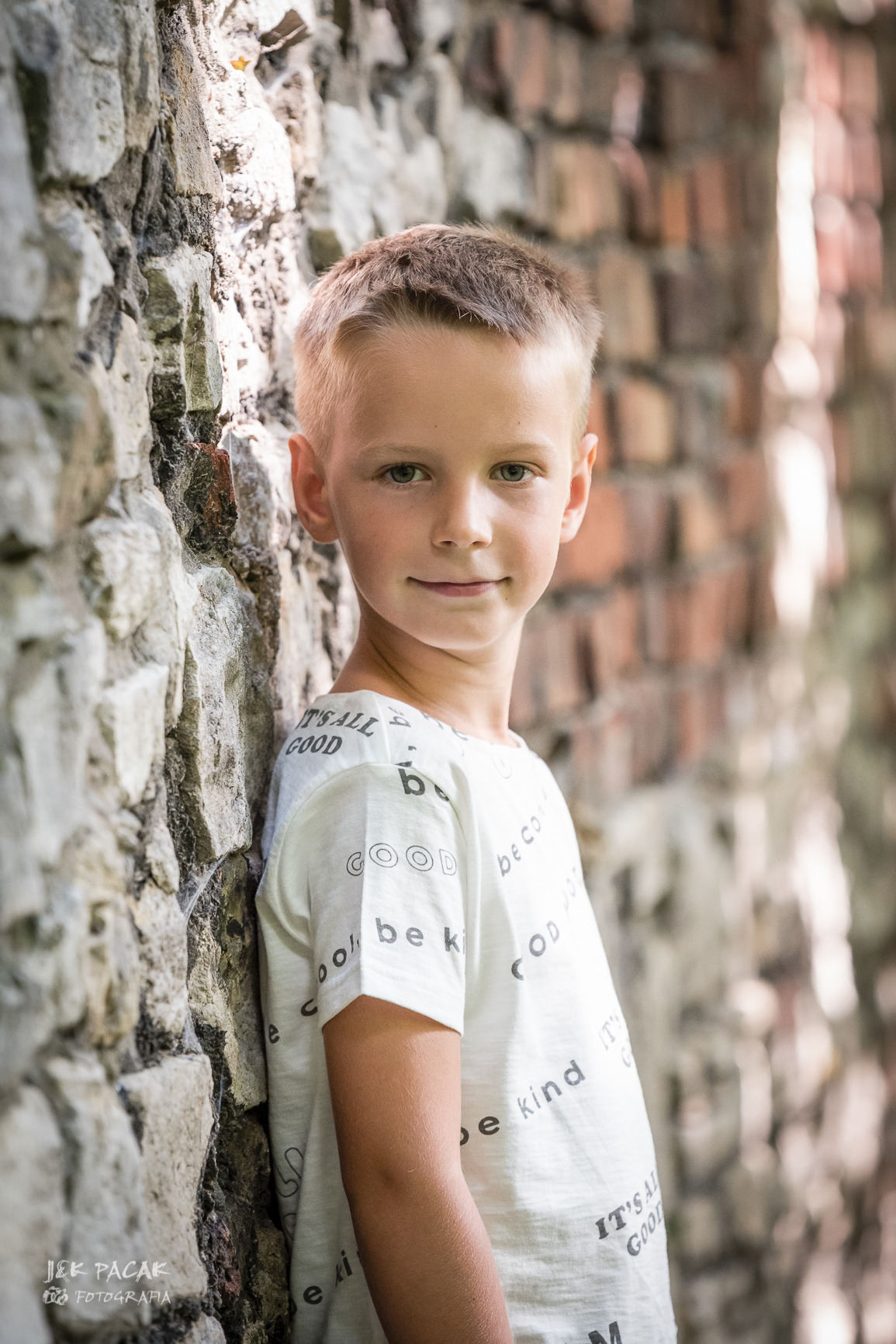 Portret uśmiechniętego chłopca opierającego się o kamienną ścianę z ceglanym murem w tle, ubrany w białą koszulkę z napisem.