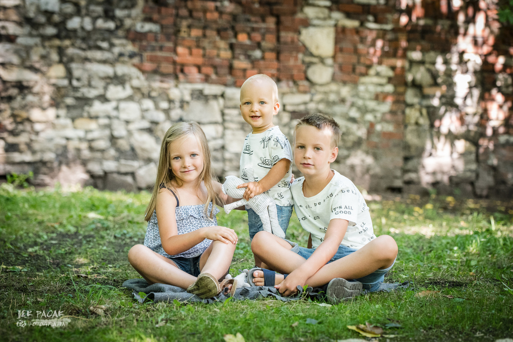 Trójka uśmiechniętych dzieci siedzi na kocu na trawie przed kamienną ścianą, najmłodsze trzyma szmacianą lalkę.