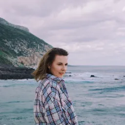 Kobieta w kraciastej marynarce spogląda przez ramię na tle morza i klifu.