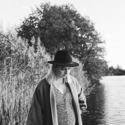 Aleksandra Jurgowiak - Czarno-białe zdjęcie kobiety w ciąży, w kapeluszu i wzorzystej sukience, stojącej nad brzegiem jeziora, z trzcinami i drzewami w tle.