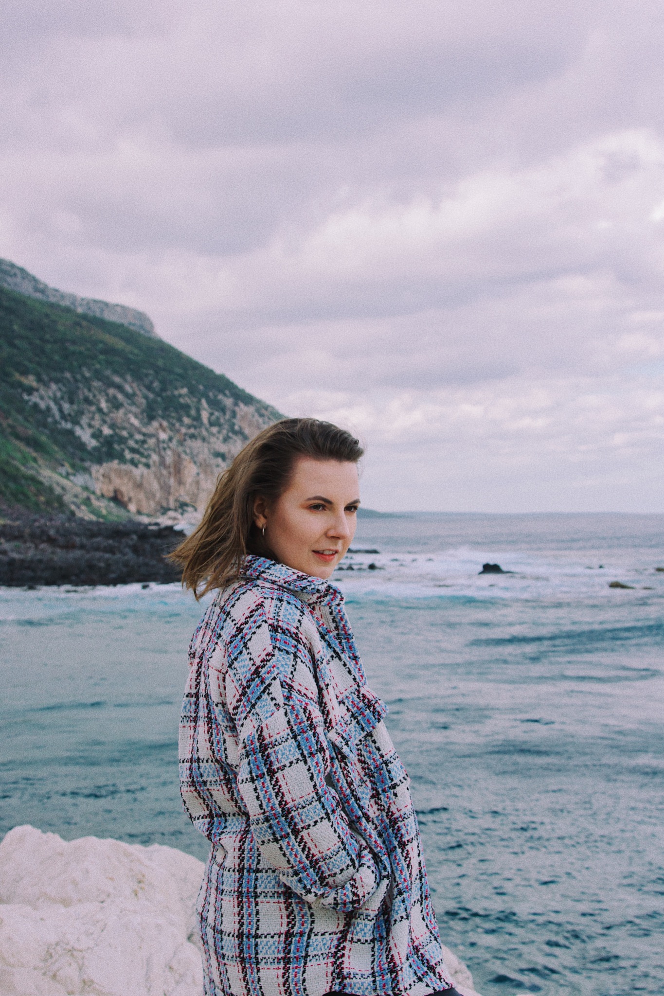 Kobieta w kraciastej marynarce spogląda przez ramię na tle morza i klifu.