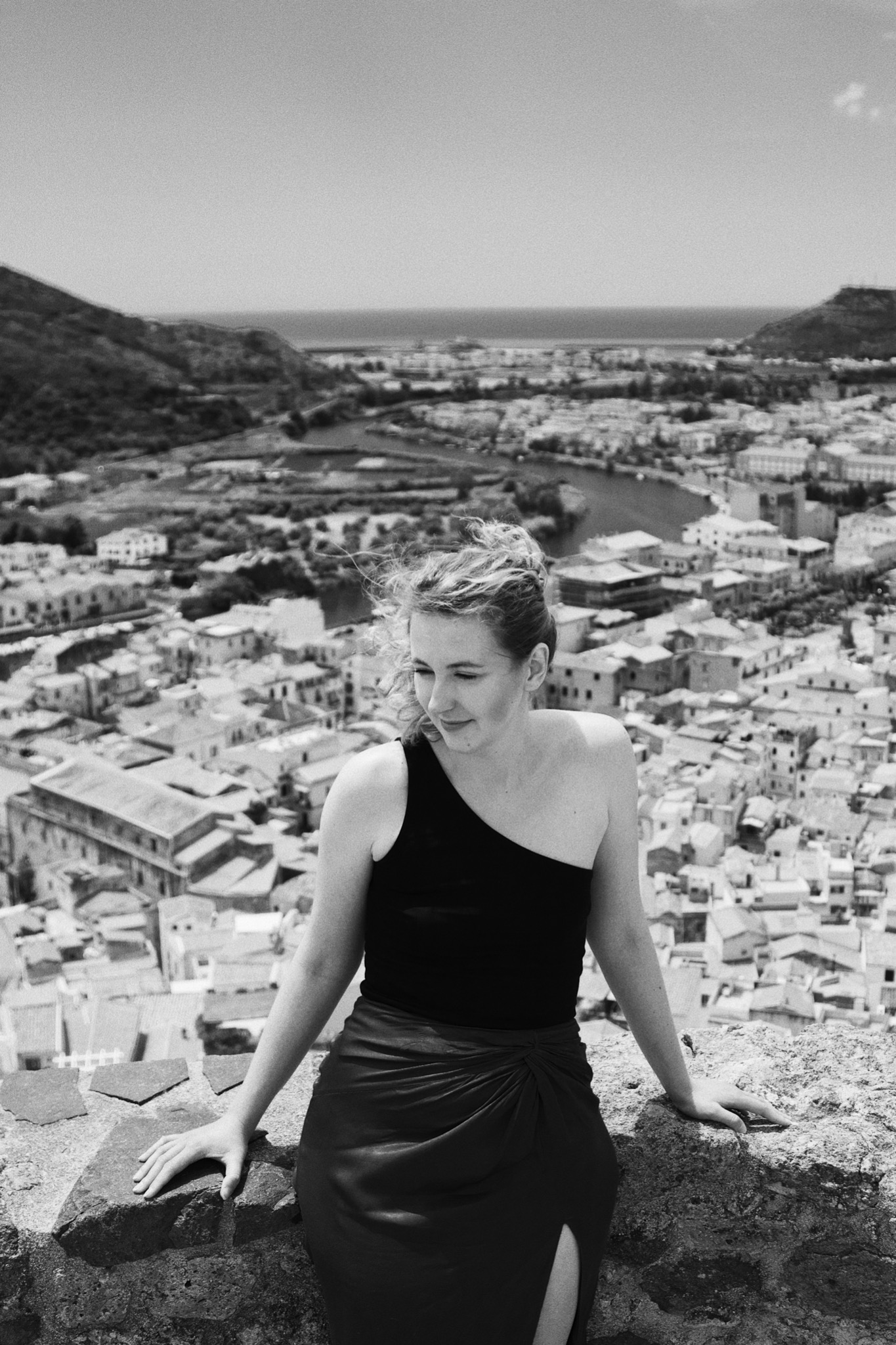 Czarno-białe zdjęcie kobiety w czarnym topie i spódnicy z rozcięciem, opierającej się o kamienny mur na tle panoramy miasta i morza.