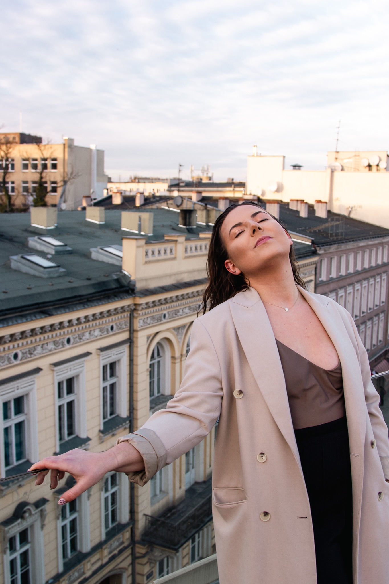 Kobieta w beżowym płaszczu, z zamkniętymi oczami, stojąca na dachu z widokiem na kamienice w stylu secesyjnym, delikatny makijaż i subtelna biżuteria.