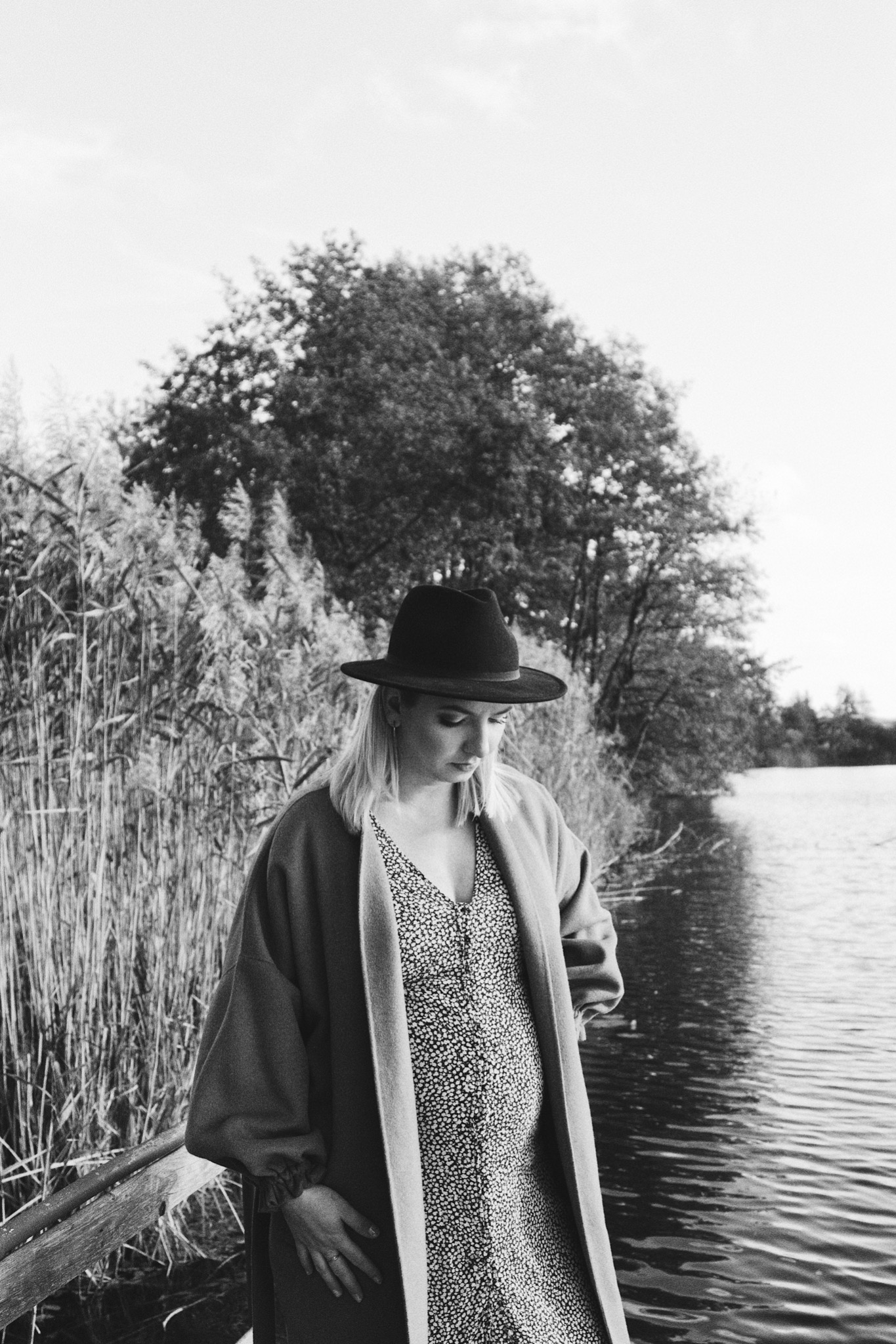 Czarno-białe zdjęcie kobiety w ciąży, w kapeluszu i wzorzystej sukience, stojącej nad brzegiem jeziora, z trzcinami i drzewami w tle.