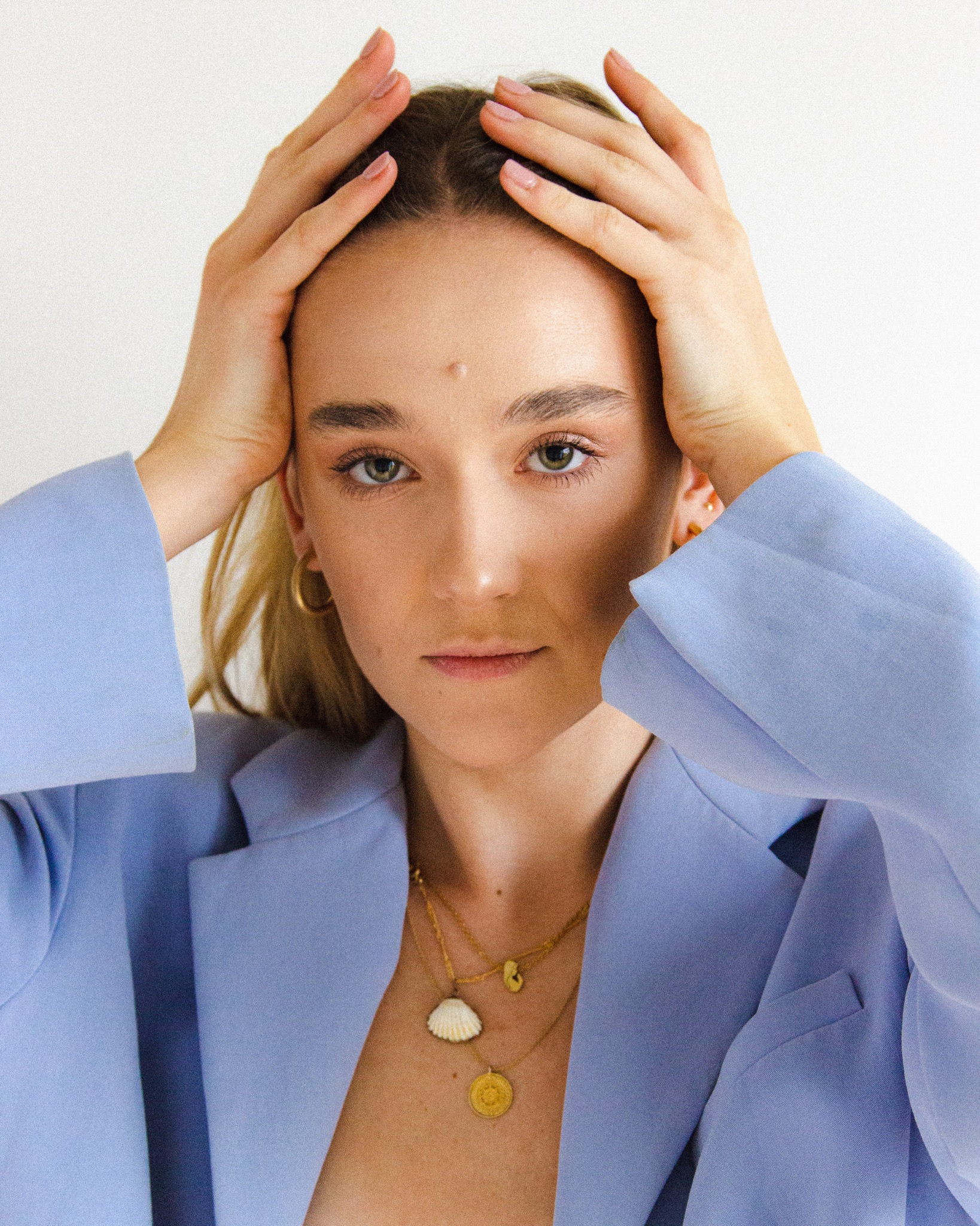 Portret kobiety w błękitnym żakiecie z rękami uniesionymi do góry, z widocznymi złotymi kolczykami i naszyjnikami na tle białej ściany.