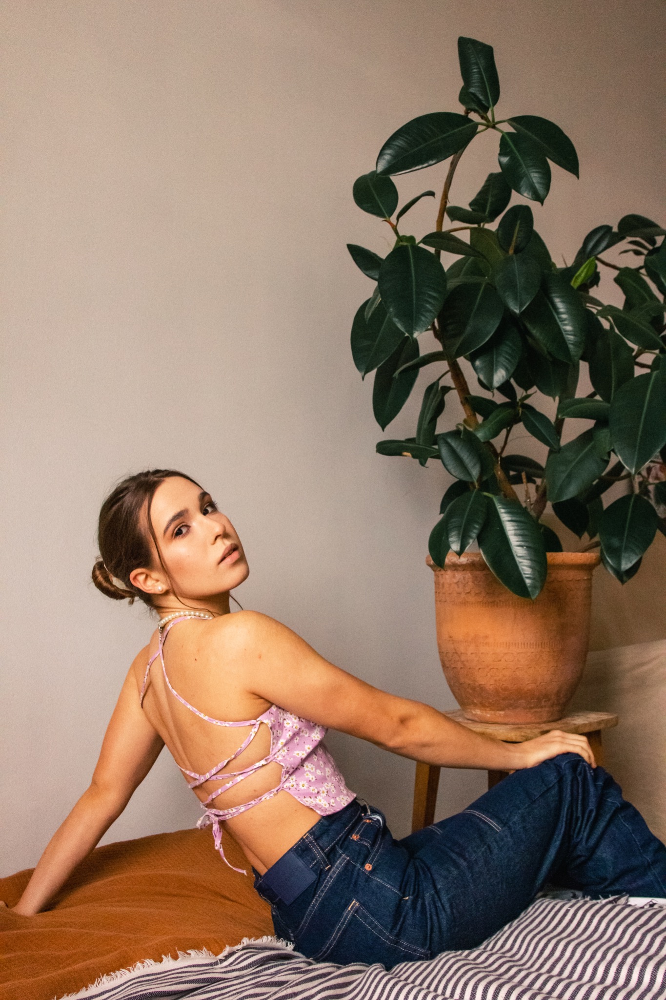 Młoda kobieta w kwiecistym topie z odkrytymi plecami i szerokich dżinsach, siedząca na łóżku obok dużej rośliny doniczkowej, spogląda w górę