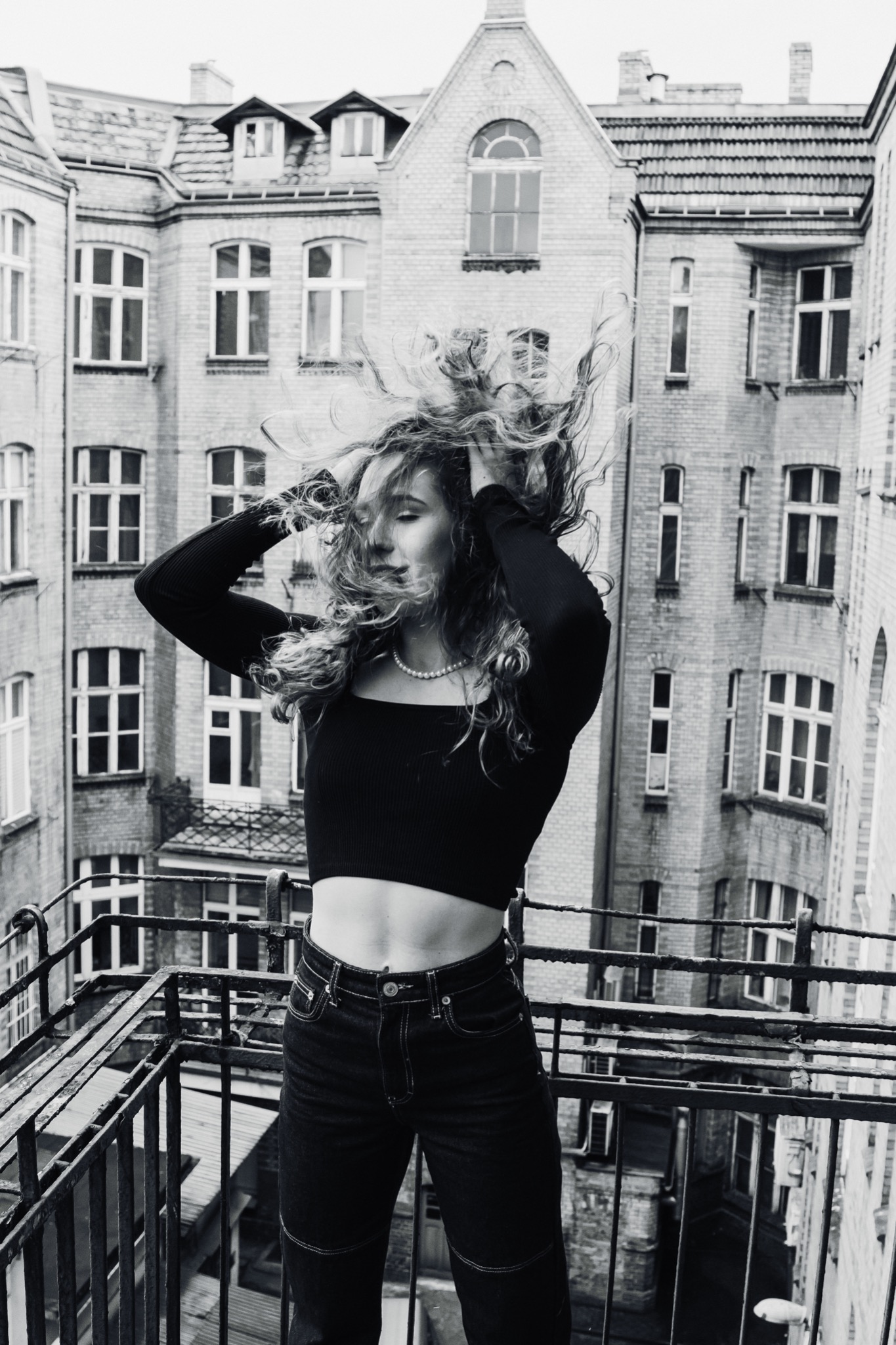 Kobieta na balkonie, w czarnym topie i dżinsach, z rozpuszczonymi włosami w ruchu, na tle kamienicy w stylu miejskim, czarno-białe zdjęcie.