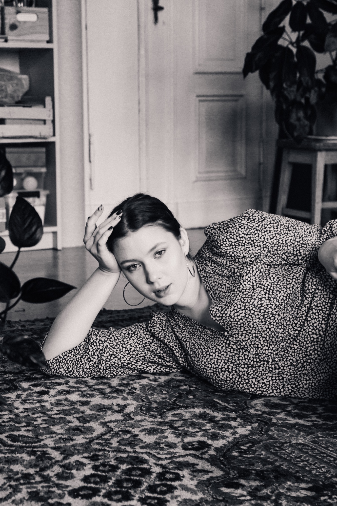 Czarno-białe zdjęcie kobiety leżącej na wzorzystym dywanie, opierającej głowę na dłoni, z widocznymi kolczykami kołami i wzorzystą sukienką, w tle regał i roślina doniczkowa.