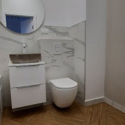 ZPP Usługi Remontowo-Wykończeniowe - Nowoczesna łazienka z umywalką w kształcie misy, podwieszaną toaletą i drewnopodobną podłogą ułożoną w jodełkę, wykończona płytkami imitującymi marmur.