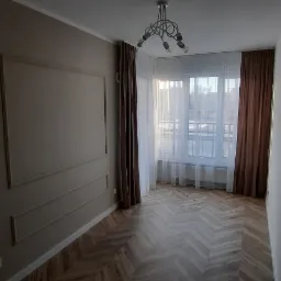 Pok&oacute;j po remoncie z parkietem ułożonym w jodełkę, beżowe ściany z dekoracyjnymi listwami, okno z białymi firanami i brązowymi zasłonami oraz nowoczesna lampa sufitowa.