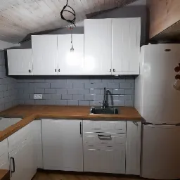 Nowoczesna kuchnia z białymi szafkami i szarymi płytkami na ścianie, drewniany blat, widoczny fragment drewnianego sufitu i ceglanej ściany.