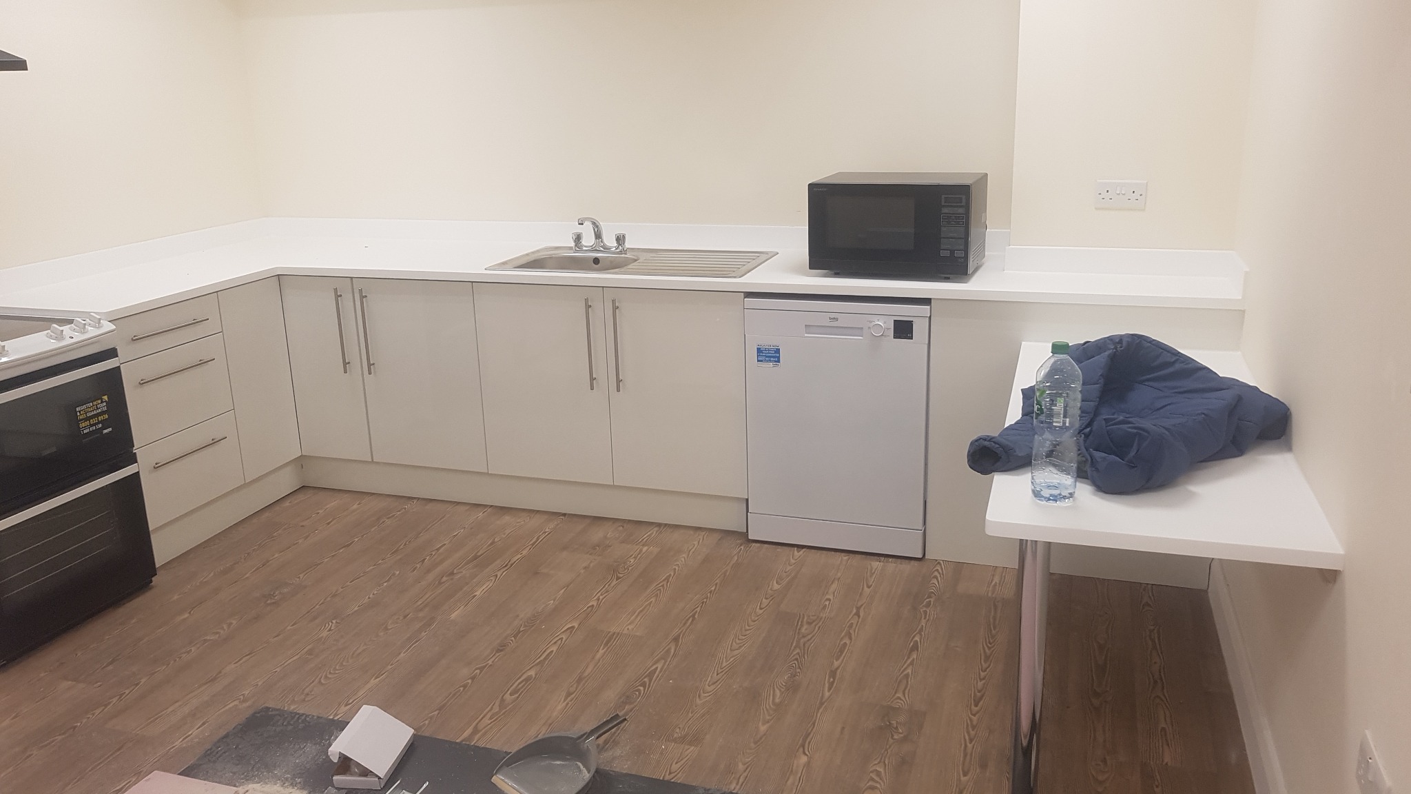 Nowoczesna kuchnia z białymi szafkami, drewnopodobną podłogą, kuchenką, mikrofalówką i zlewem. Na blacie leży niebieska kurtka i butelka wody.