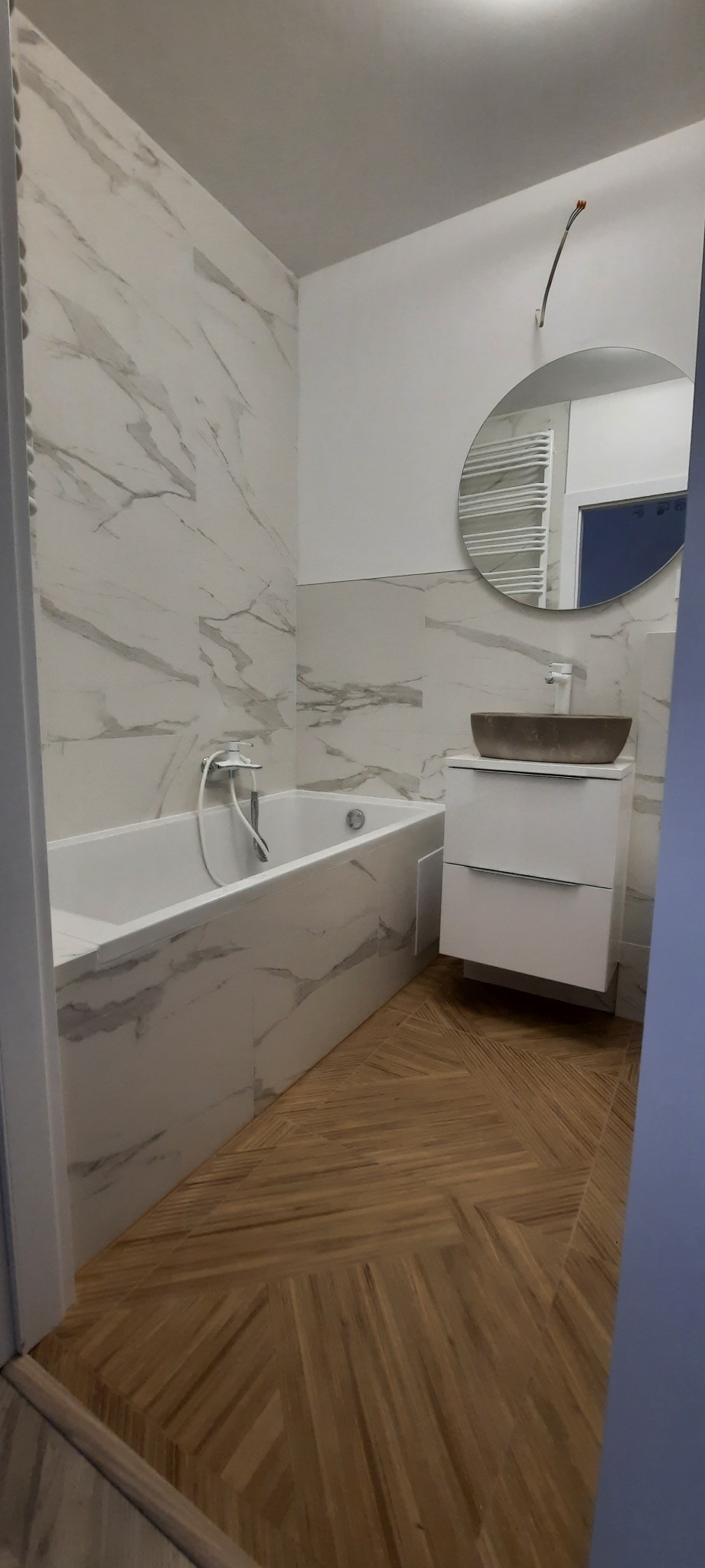 Nowoczesna łazienka z marmurowymi płytkami, drewnianą podłogą w jodełkę, okrągłym lustrem i umywalką w odcieniach brązu na białej szafce.