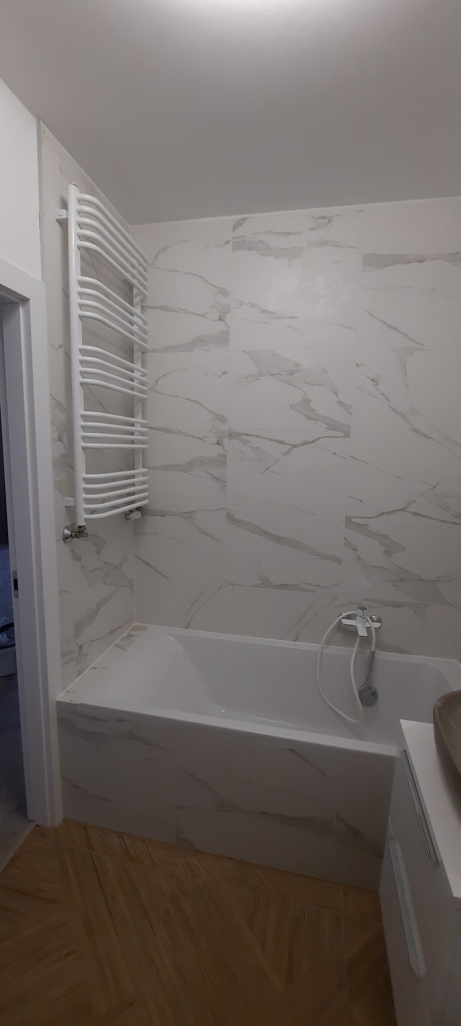 Nowoczesna łazienka z wanną obłożoną marmurową płytką, grzejnikiem drabinkowym i parkietem ułożonym w jodełkę.