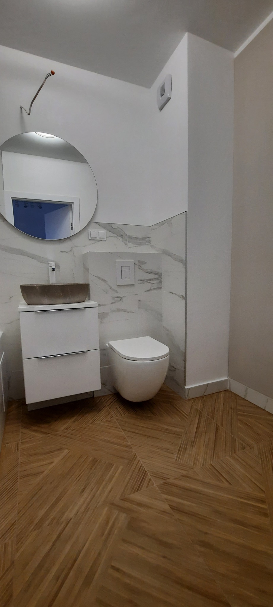 Nowoczesna łazienka z umywalką w kształcie misy, podwieszaną toaletą i drewnopodobną podłogą ułożoną w jodełkę, wykończona płytkami imitującymi marmur.