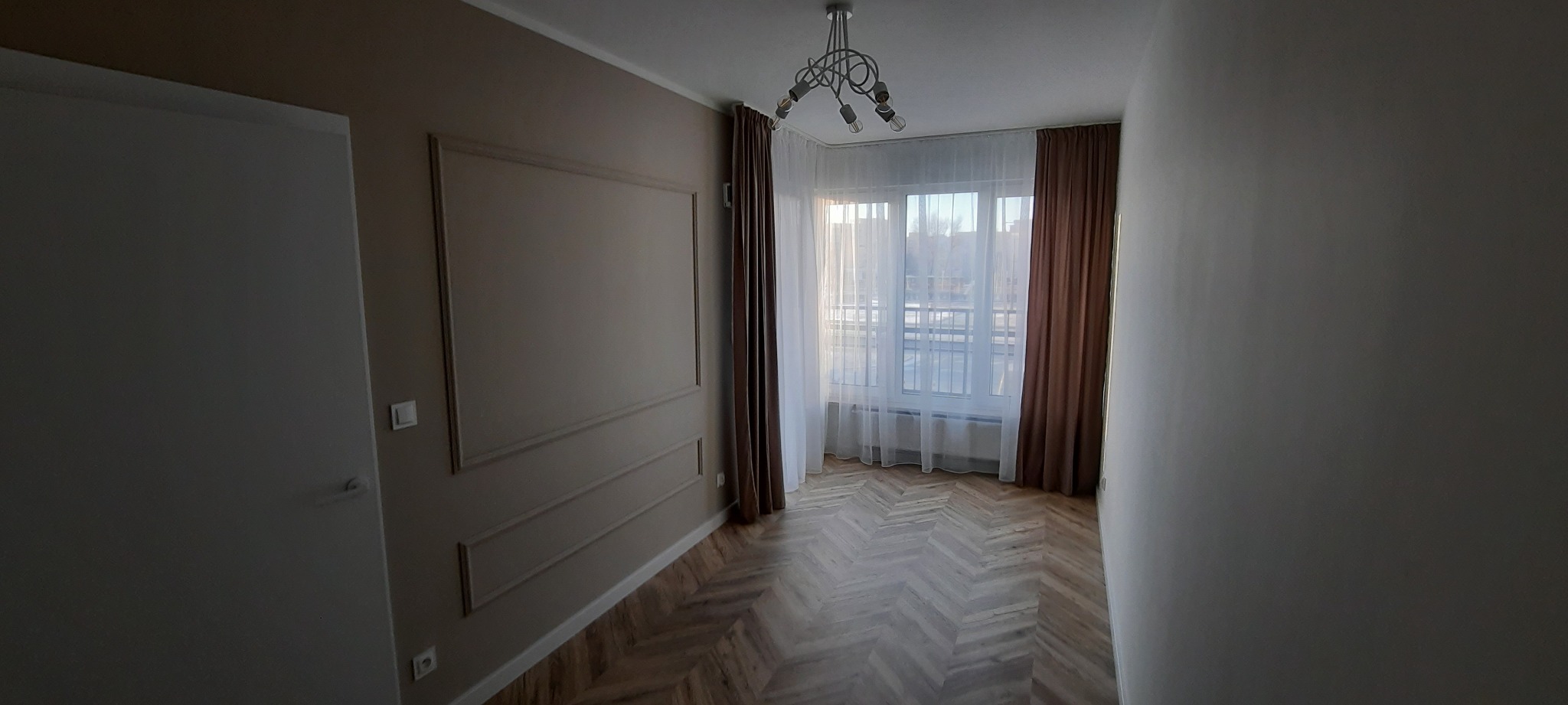 Pokój po remoncie z parkietem ułożonym w jodełkę, beżowe ściany z dekoracyjnymi listwami, okno z białymi firanami i brązowymi zasłonami oraz nowoczesna lampa sufitowa.